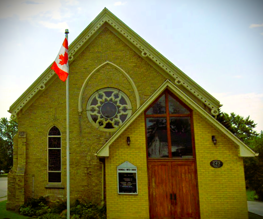 a church building with yellow bricks and wood doors with a canada flag in front in the background a blue sky and trees
