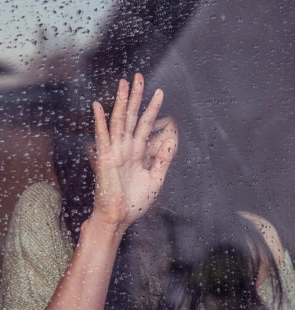 rain on a window in the background a woman with her hand up on the window
