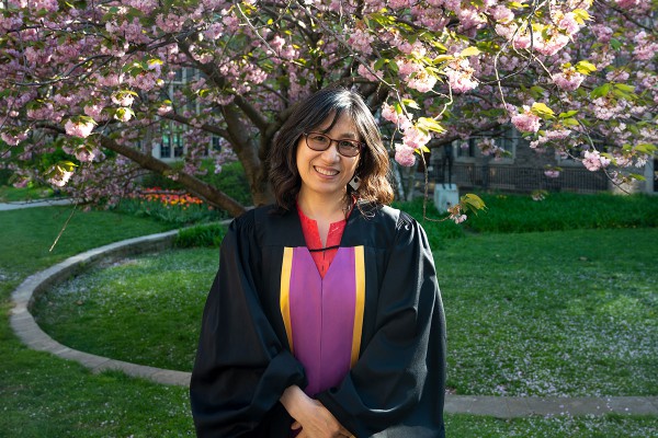 a woman with shoulder length brown hair wearing glasses and smiling, she is wearing a black robe with a purple and yellow and pink dress underneath in the background is a blooming magnolia tree