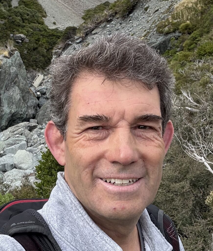 a man with brown hair smiling at the camera wearing a grey sweatshirt and black backpack in the background are mossy rocks