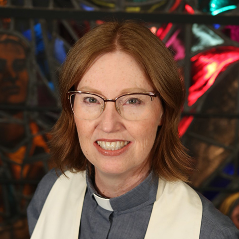 a woman with shoulder length red hair, wearing glasses and smiling, wearing an alb and a clergy collar in the backgroun is red and blue stained glass